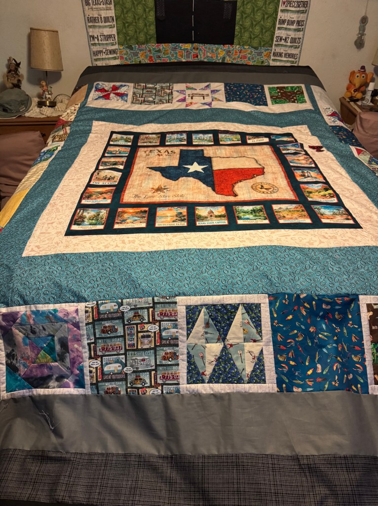 A close-up view of a quilt featuring a Texas map design, surrounded by various colorful patches and patterns. The quilt is displayed on a bed, accompanied by a bedside lamp and other bedroom decor.