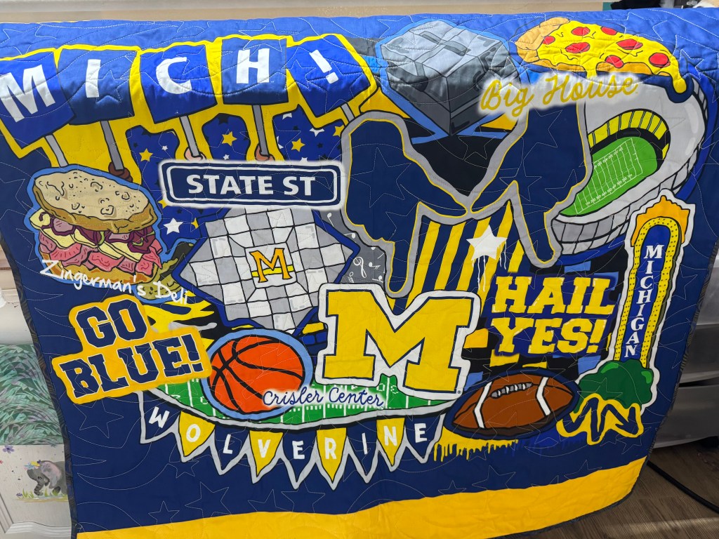 A colorful quilt featuring various iconic elements related to the University of Michigan, including the school's logo, football, basketball, and local food items like a sandwich and pizza. The design includes phrases like 'Go Blue!' and 'Hail Yes!' against a blue and yellow background.