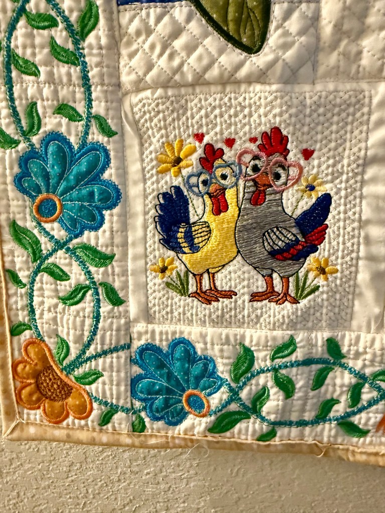 A colorful quilted fabric featuring an embroidered design of two cartoon chickens, one yellow and one gray, surrounded by flowers and green vines.