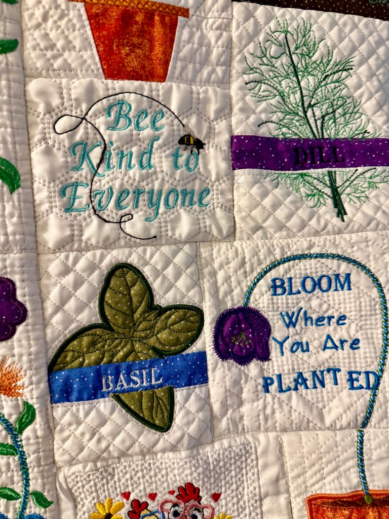 A quilt featuring embroidered squares with phrases and images, including 'Bee Kind to Everyone', 'BASIL', and 'BLOOM Where You Are PLANTED', alongside decorative floral and herb designs.